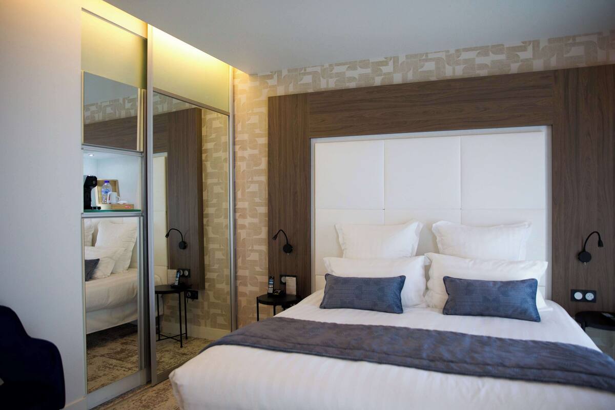 A modern hotel room with a large bed, a bedside table, a mirror, and a wardrobe.