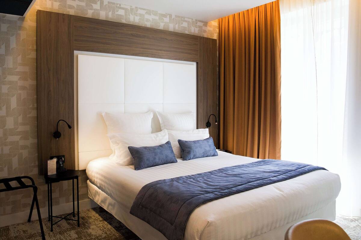 A neatly made bed with a white headboard, two pillows, and a blue throw blanket. There is a small table with a lamp beside the bed.