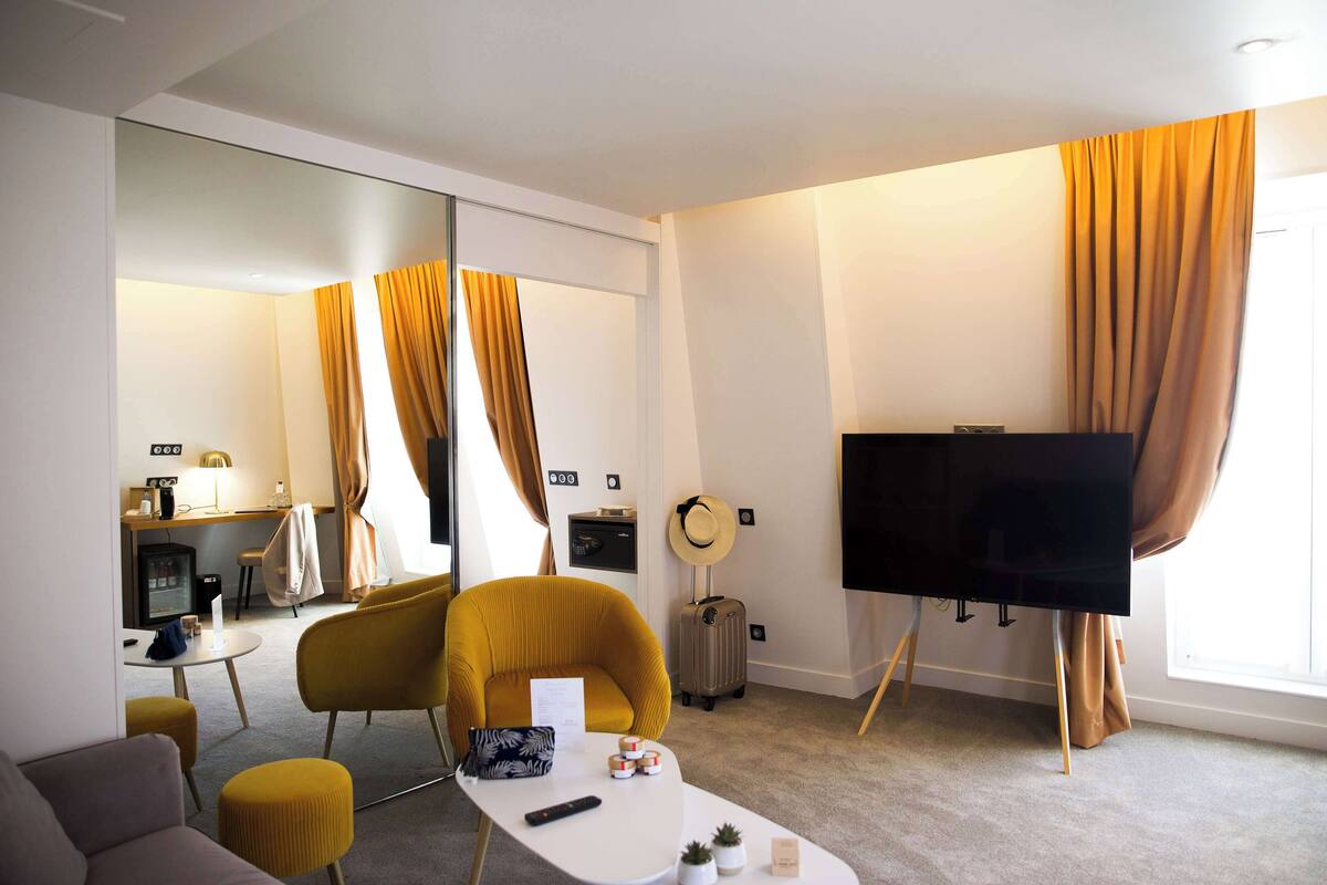 A hotel room with a sofa, a small table with a remote control, a television, a mirror, and a window with curtains.