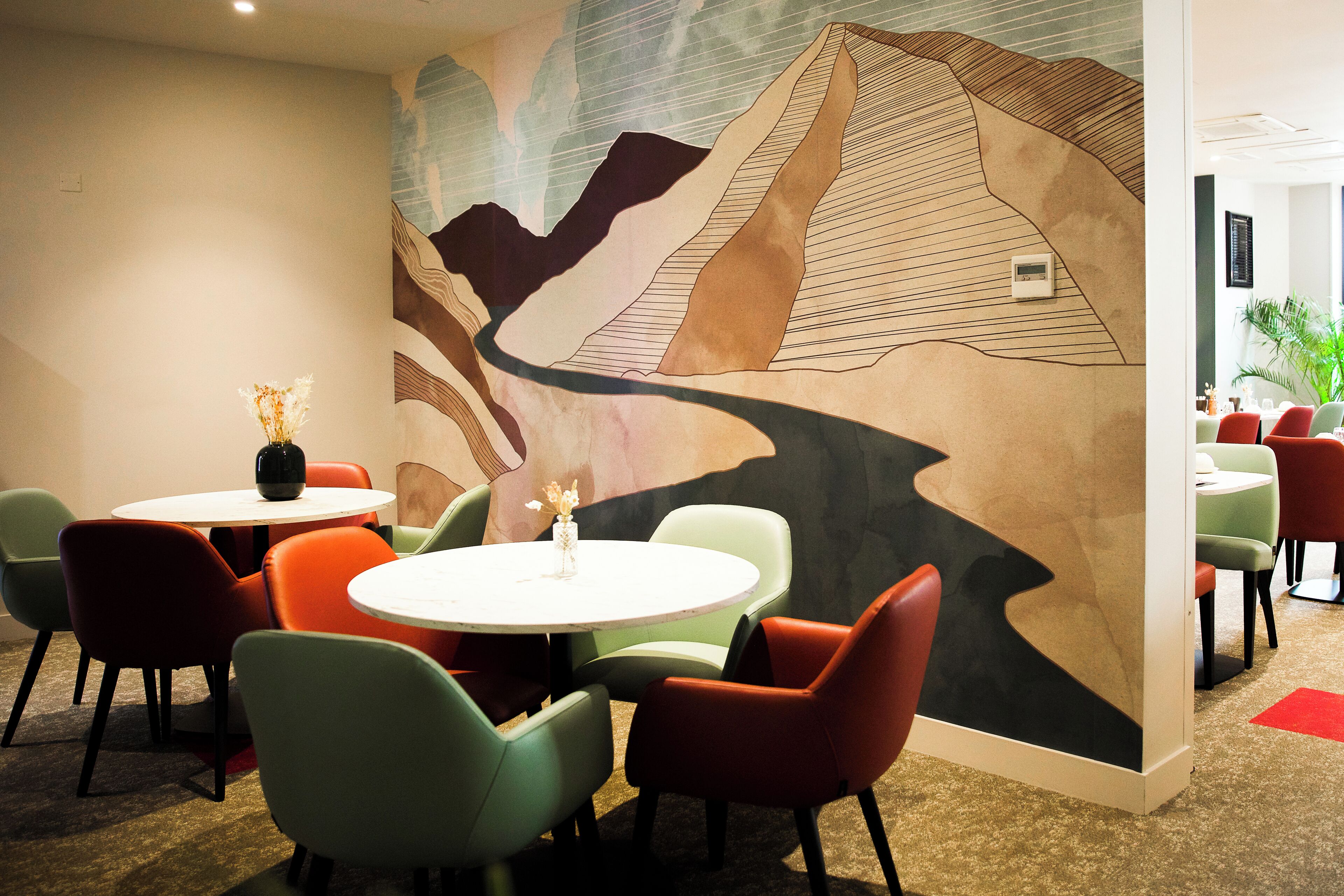 A modern conference room with round tables and colorful chairs, a large abstract mural on the wall, and a small plant on one table.