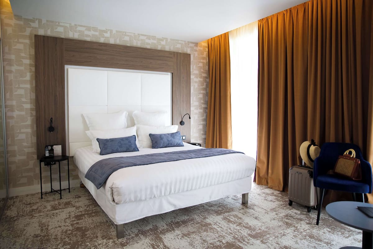 A hotel room with a large bed, a chair, a suitcase, and a small table.