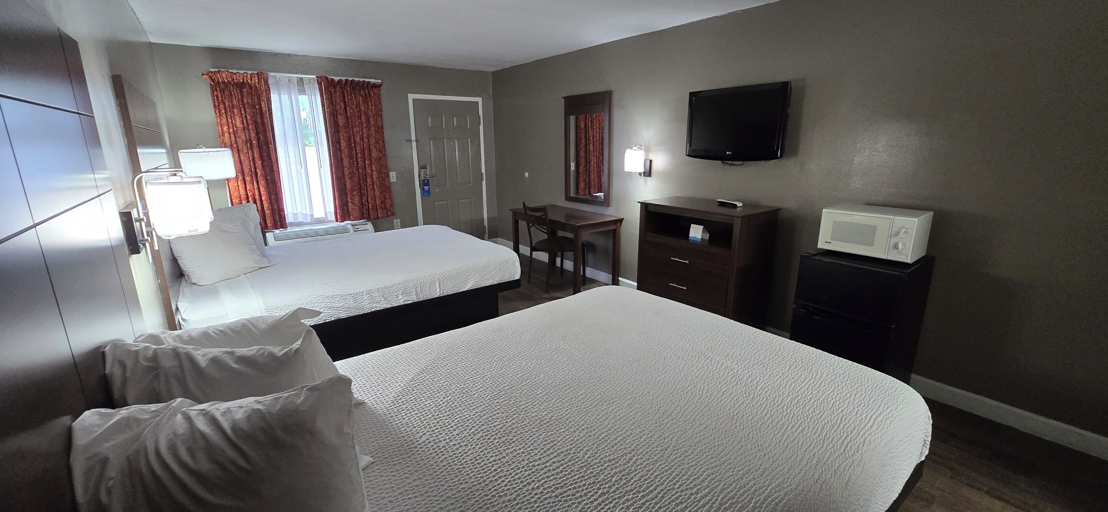 A hotel room with two beds, a desk, a TV, a microwave, and a window with curtains.