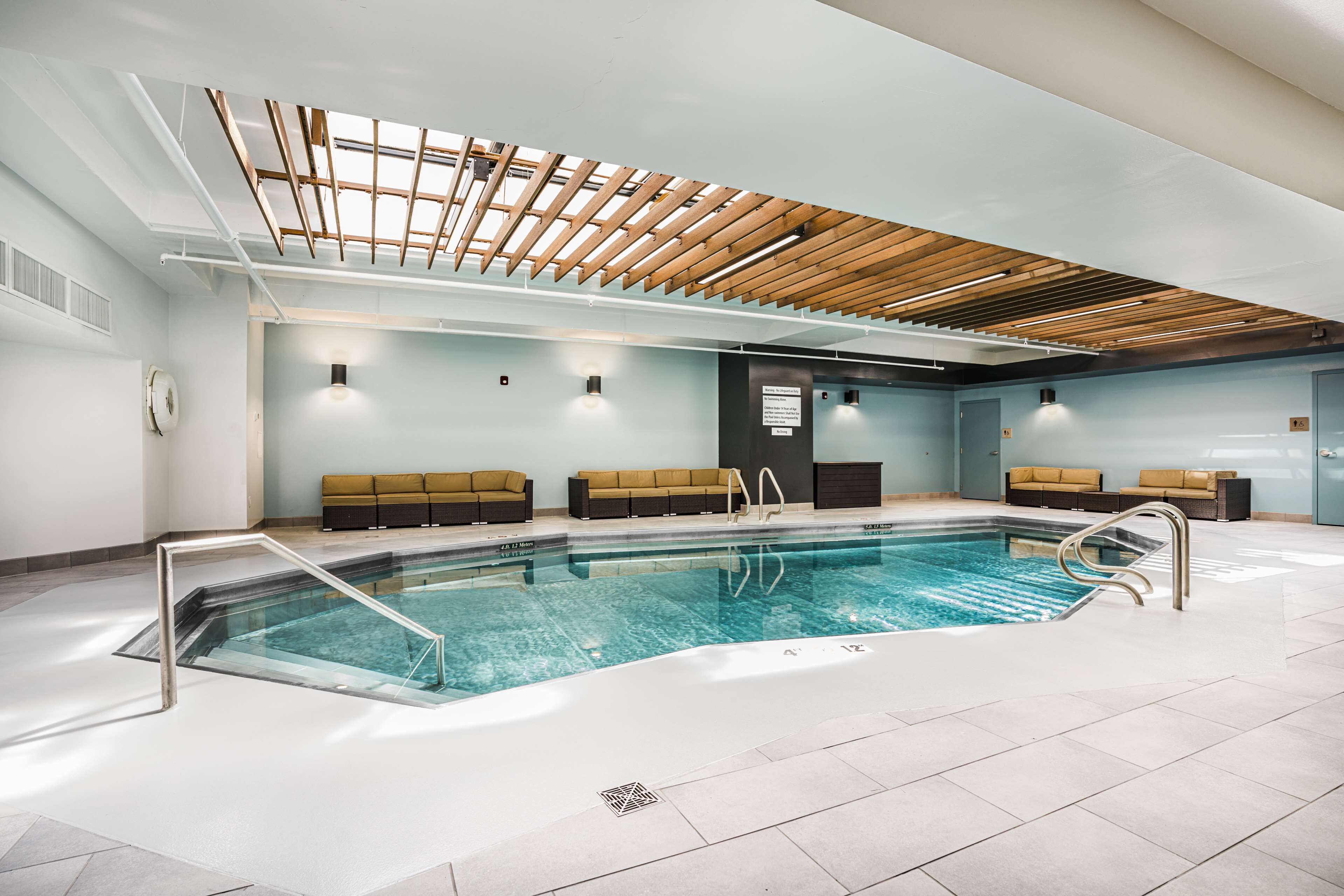 Una piscina interna con soffitto in legno, pareti azzurro chiaro e zone relax color beige.