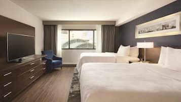 A hotel room with two beds, a TV, a desk, and a window with a view of buildings.