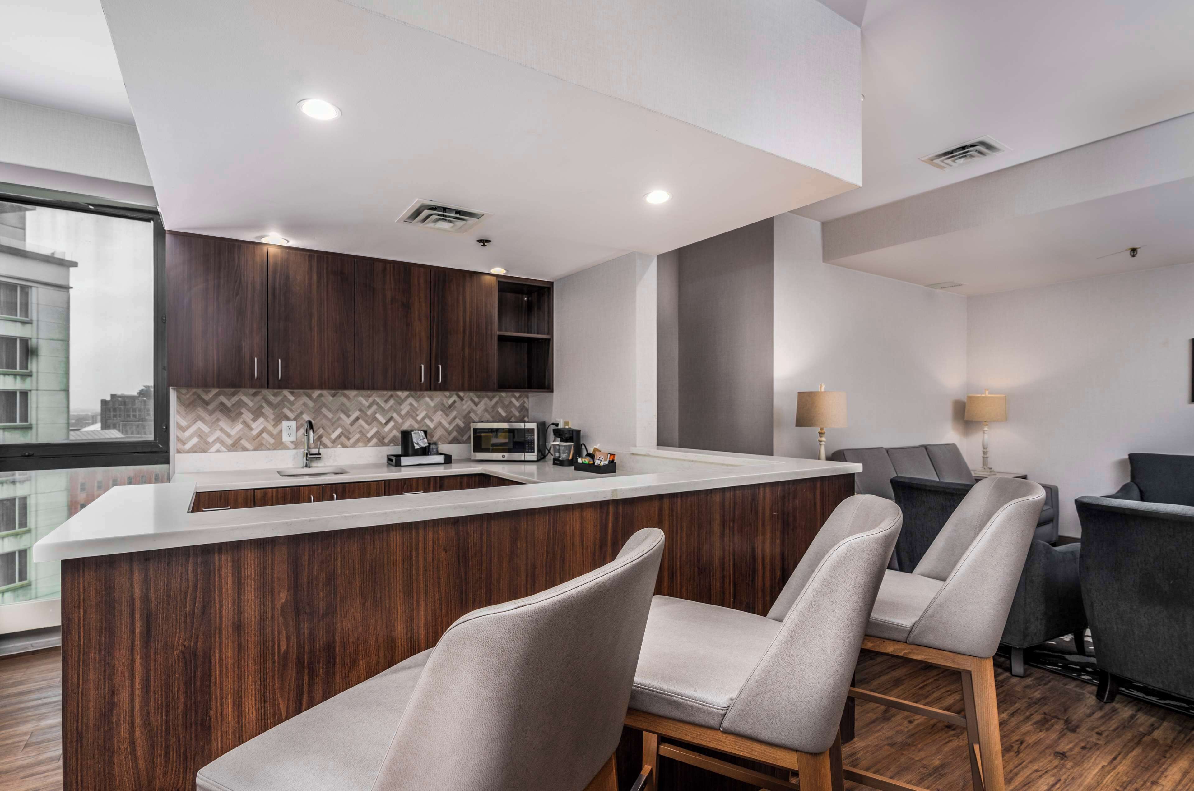 A modern kitchen with a central island, dark wood cabinets, and a herringbone backsplash. There are two bar stools in front of the island and a dining area with a dark sofa and two lamps.