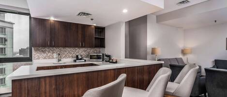 A modern kitchen with a central island, dark wood cabinets, and a herringbone backsplash. There are two bar stools in front of the island and a dining area with a dark sofa and two lamps.