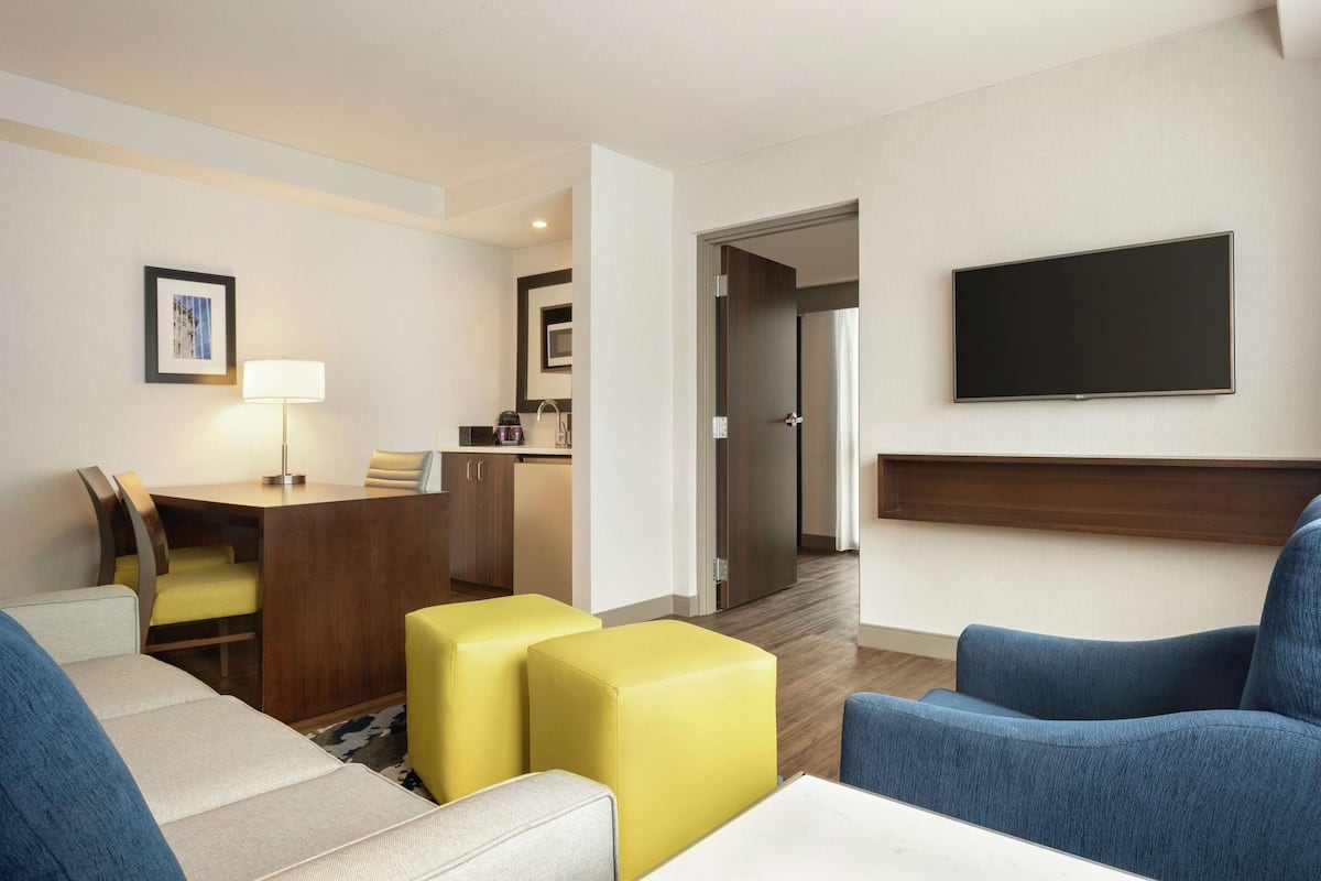 A hotel room with a sofa, yellow ottoman, a flat-screen TV mounted on the wall, a desk with a lamp, and a kitchenette in the background.