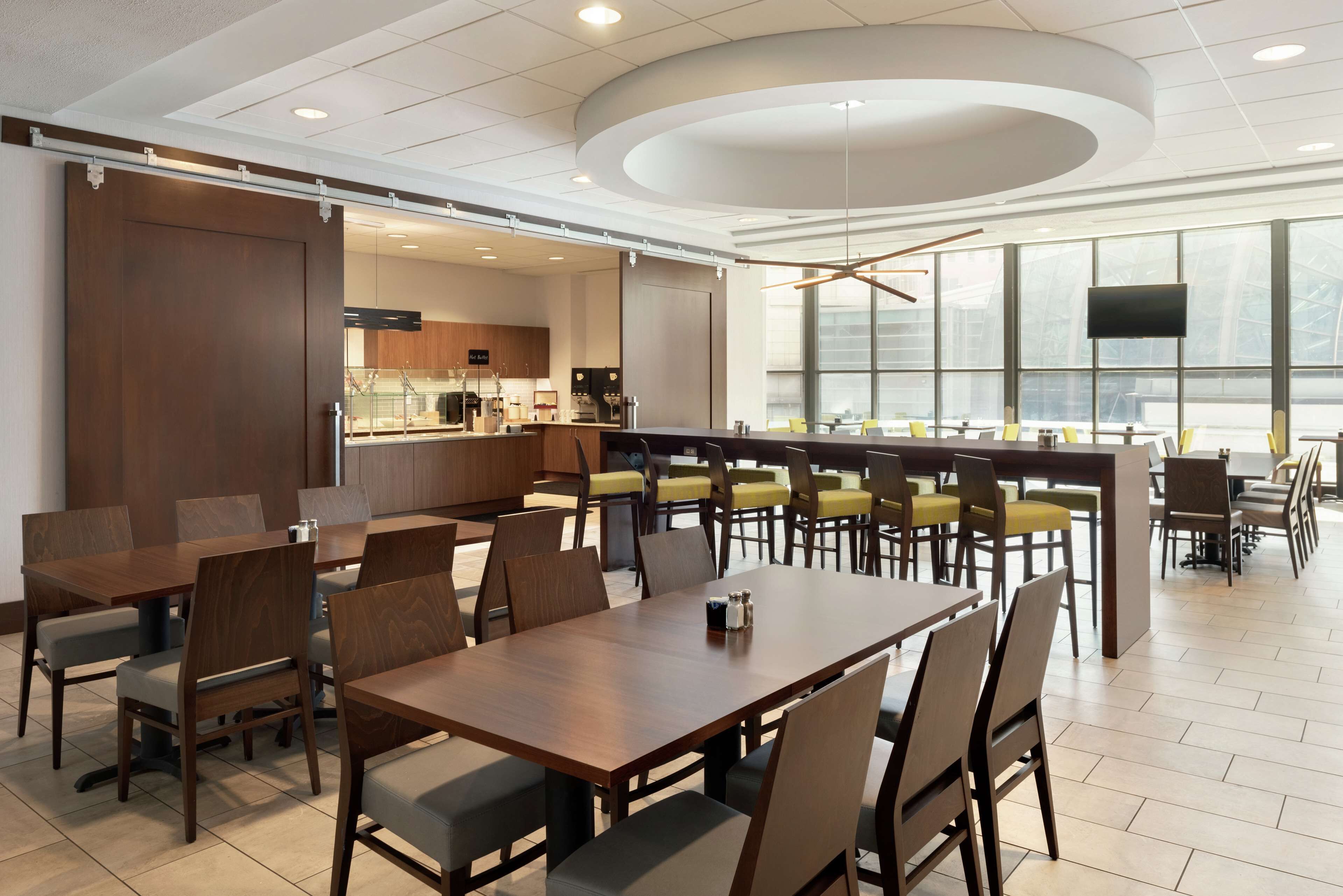 A modern dining area with wooden tables and chairs, a kitchen in the background, and large windows providing natural light.