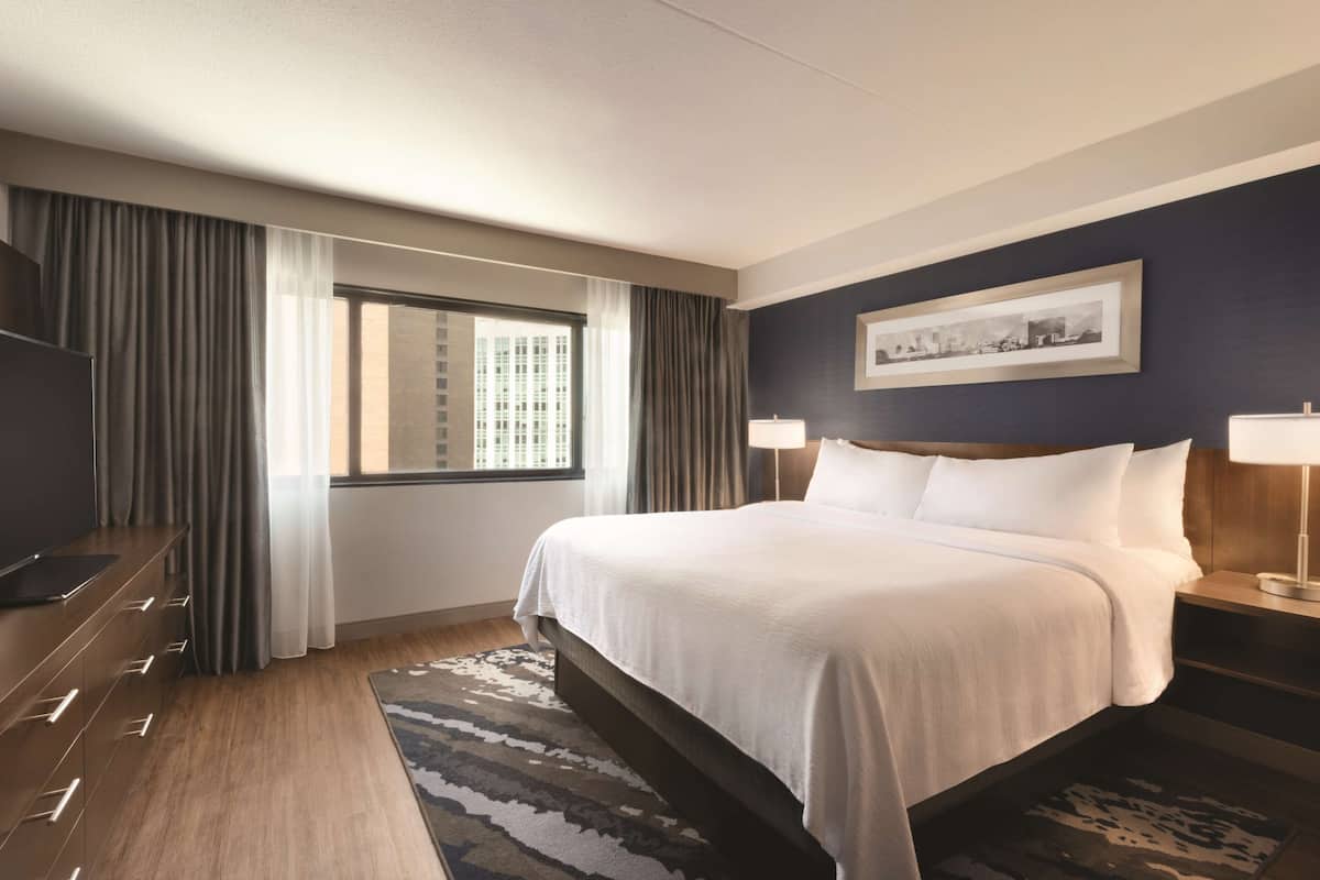 A modern hotel room with a large bed, a flat-screen TV, a desk, and a window with curtains.