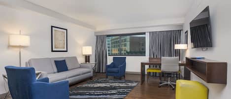 Ein modernes Hotelzimmer mit grauem Sofa, blauem Sessel, gelbem Hocker und einer Essecke mit Tisch und Stühlen.