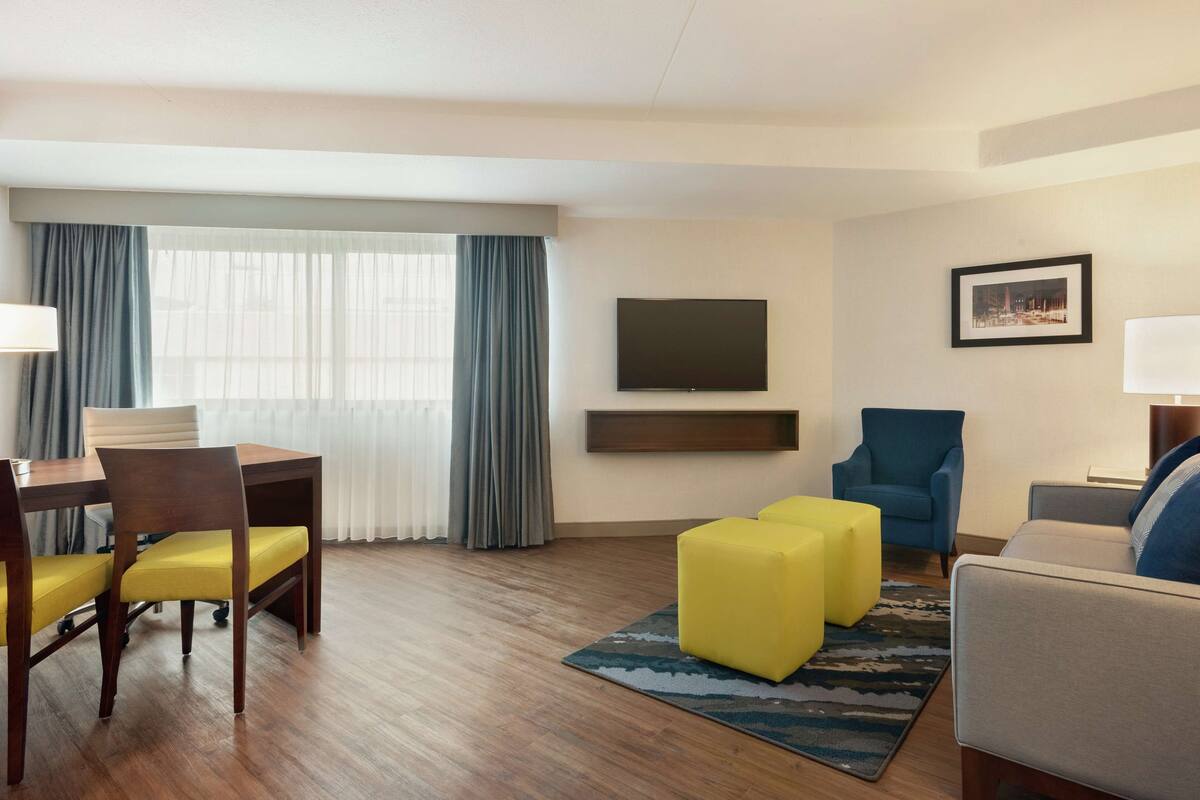 A hotel room with a wooden floor, a dining table with yellow chairs, a blue armchair, a gray sofa, a flat-screen TV, and a framed picture on the wall.