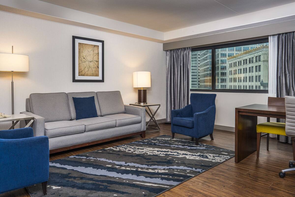 A modern hotel room with a grey sofa, blue armchairs, a wooden desk, and a large window with curtains.
