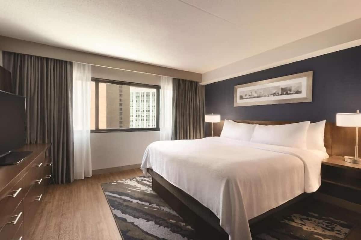 A hotel room with a bed, a TV, a desk, a window with curtains, and a framed picture on the wall.