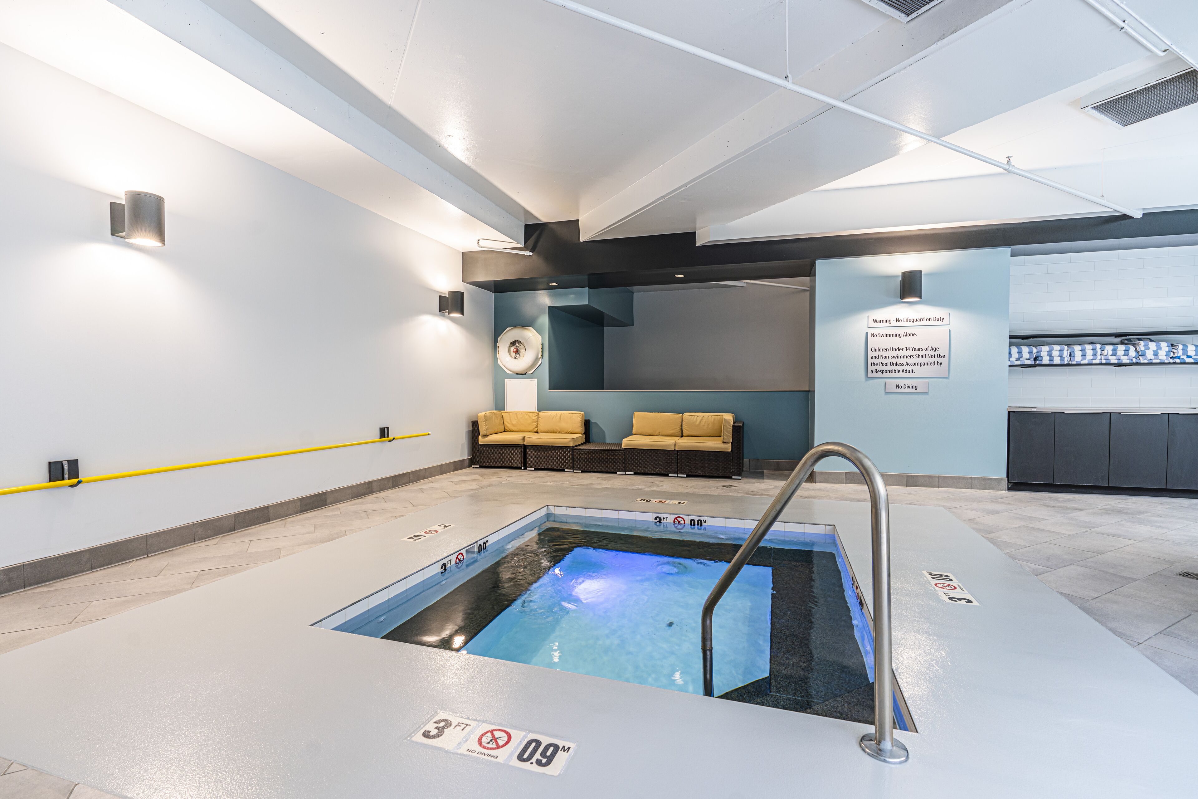 A modern indoor pool area with a hot tub, lounge chairs, and a safety sign.