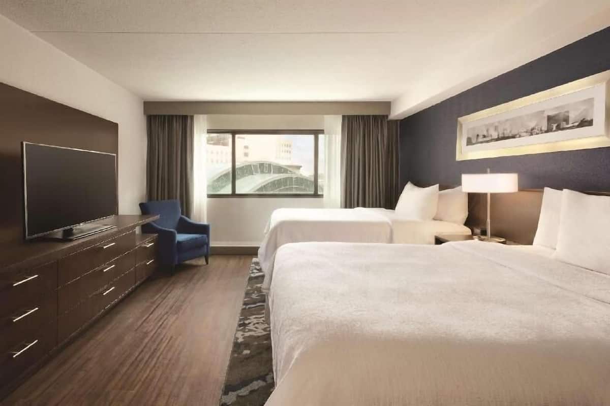 A hotel room with two beds, a TV, a desk, a chair, and a window with curtains.