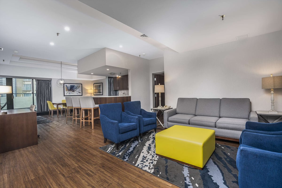 A modern living room with a grey sofa, blue armchairs, and a yellow ottoman. There is a dining area with a table and chairs, and a kitchen in the background.
