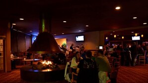 A restaurant with a central fire feature, tables, chairs, and a television screen. - Townhouse Hotel Grand Forks (Grand Forks)