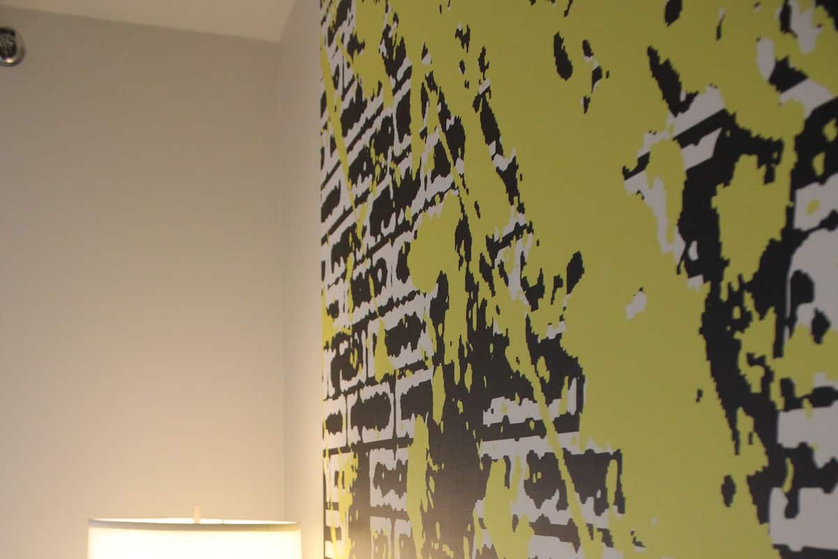 A hotel room with a bed, a nightstand, a lamp, and a wall with a yellow and black abstract design.