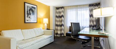 Une chambre d’hôtel avec un canapé blanc, un bureau en verre, un fauteuil de bureau noir, un tableau encadré au mur, une lampe et une fenêtre avec des rideaux à motifs.