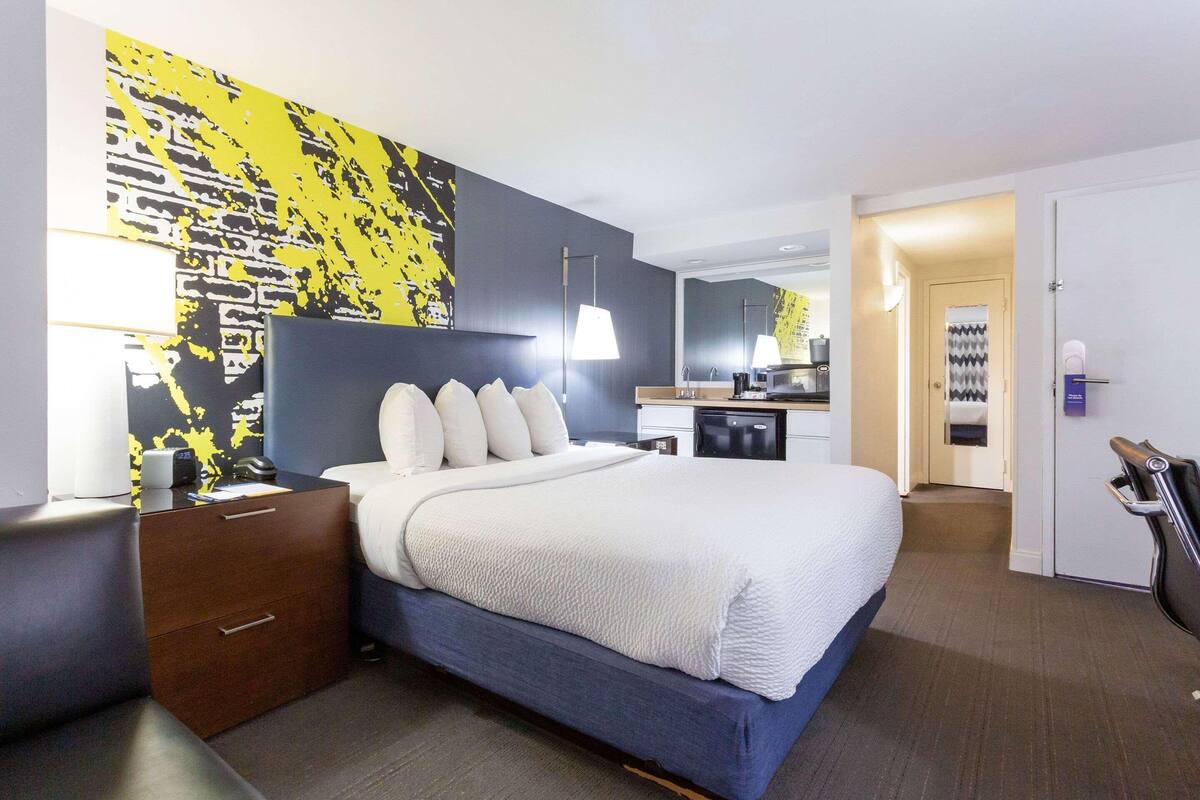 A hotel room with a bed, bedside table, lamp, and a wall with a yellow abstract design.