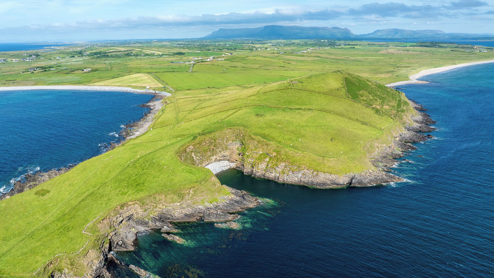 Vista aérea de un paisaje costero con campos verdes, un litoral rocoso y un mar azul cristalino.