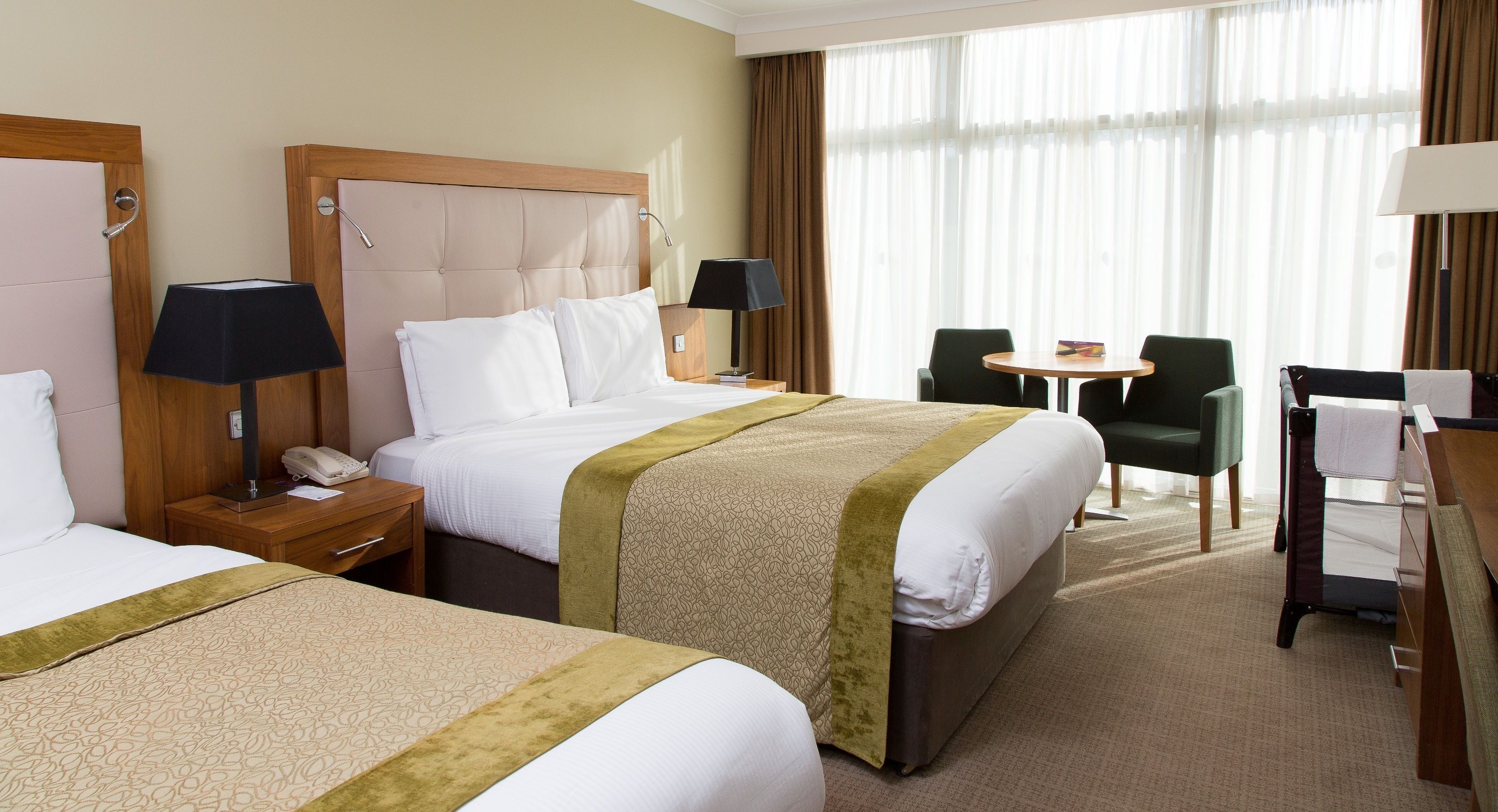 Ein Hotelzimmer mit zwei Betten, einem kleinen Tisch, zwei Stühlen, einem Schreibtisch und einem großen Fenster mit Vorhängen.