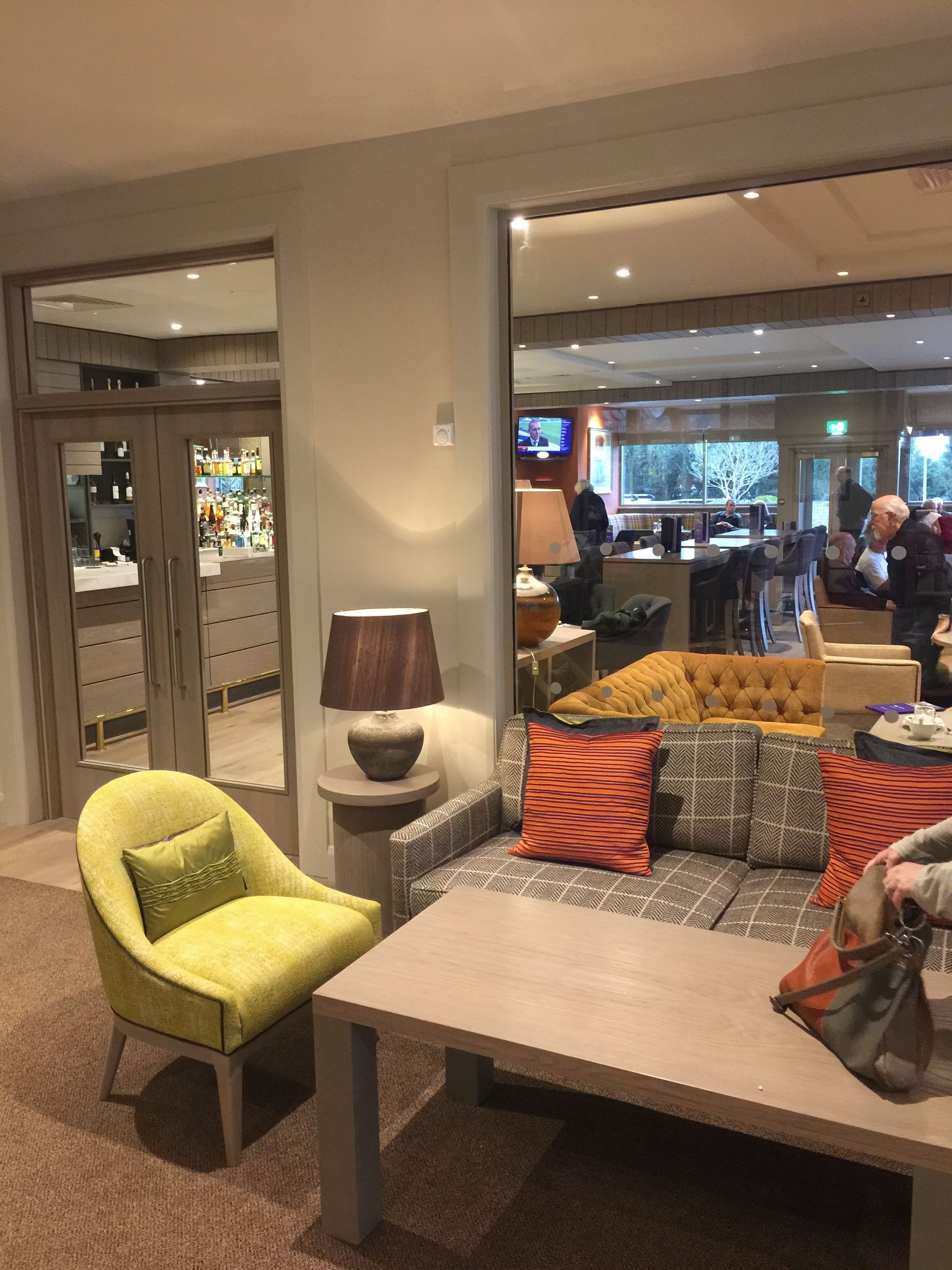 A hotel lobby with a seating area featuring a sofa and armchairs, a coffee table, and a lamp. There is a bar area visible through a glass partition.