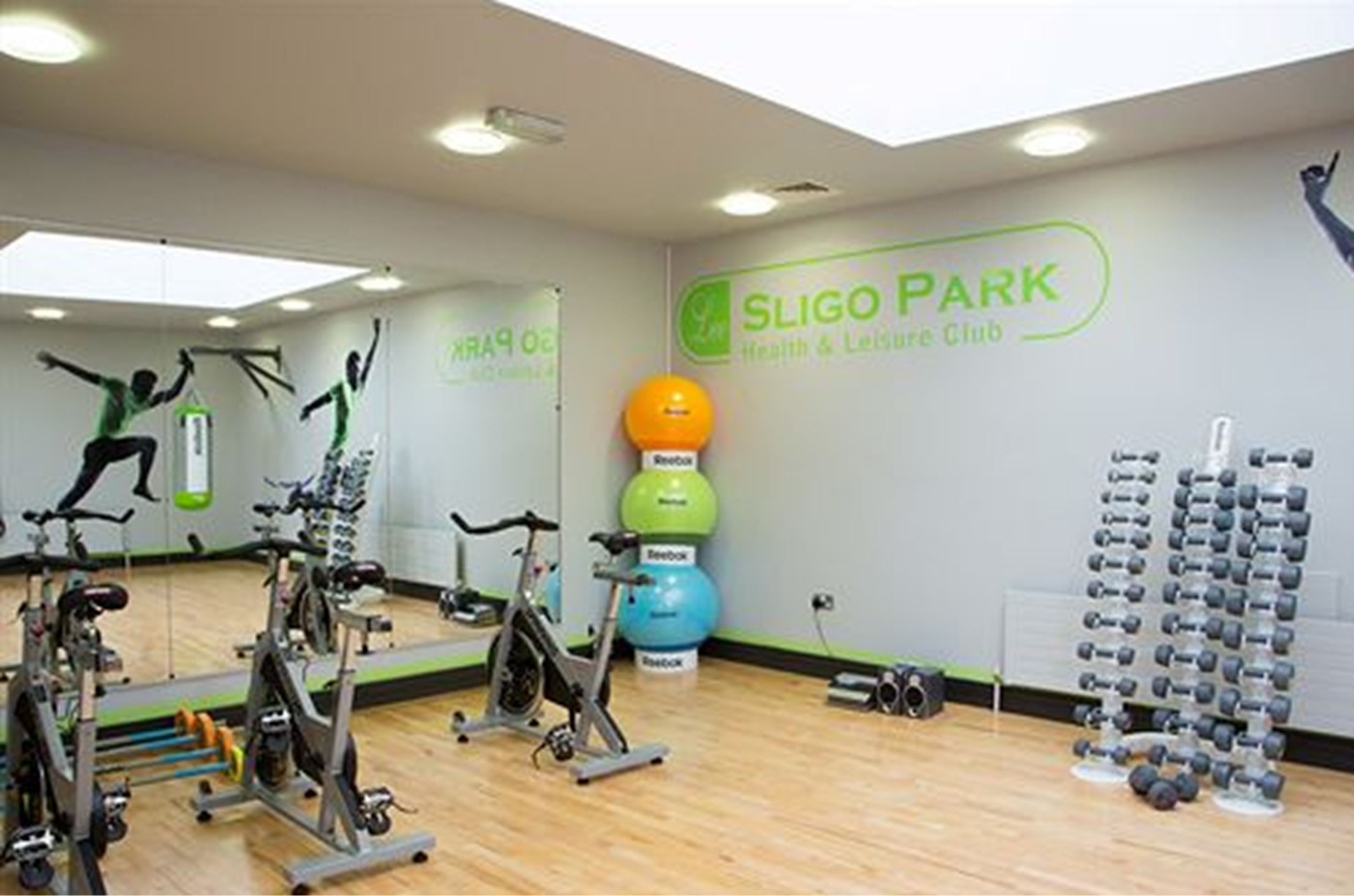 Un gimnasio con bicicletas estáticas, una pelota Reebok sujeta a la pared y una pared llena de mancuernas.