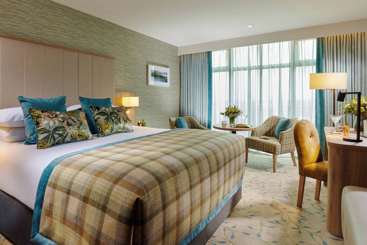 Une chambre d’hôtel avec un grand lit, deux tables de chevet, un bureau avec une chaise, une fenêtre avec des rideaux et une lampe fixée au mur.