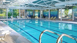 Una piscina interior con agua azul cristalina, sillas blancas junto a la piscina y un techo de azulejos con diseño geométrico.