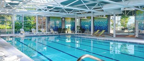 Una piscina interior con agua azul cristalina, sillas blancas junto a la piscina y un techo de azulejos con diseño geométrico.