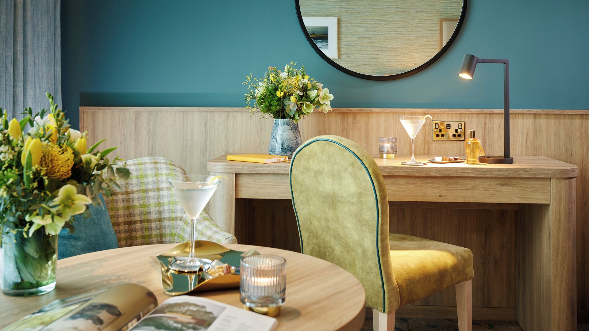 A modern room with a wooden desk, a round table, a green chair, a vase with flowers, a martini glass, and a lamp.