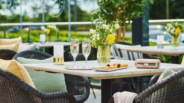 Un área de comedor al aire libre con sillas de mimbre, una mesa con un jarrón de flores, dos copas de vino blanco y un plato de postre.