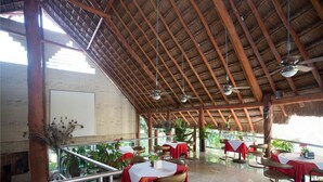 A spacious indoor dining area with high ceilings, wooden beams, and a thatched roof. Tables are set with red tablecloths, and there are potted plants and a large vase of flowers. - Hotel Plaza Caribe (Cancun)