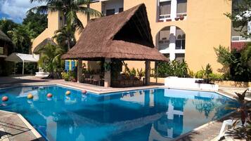 Una piscina con una estructura de techo de paja, rodeada de palmeras y un edificio con ventanas de arco.