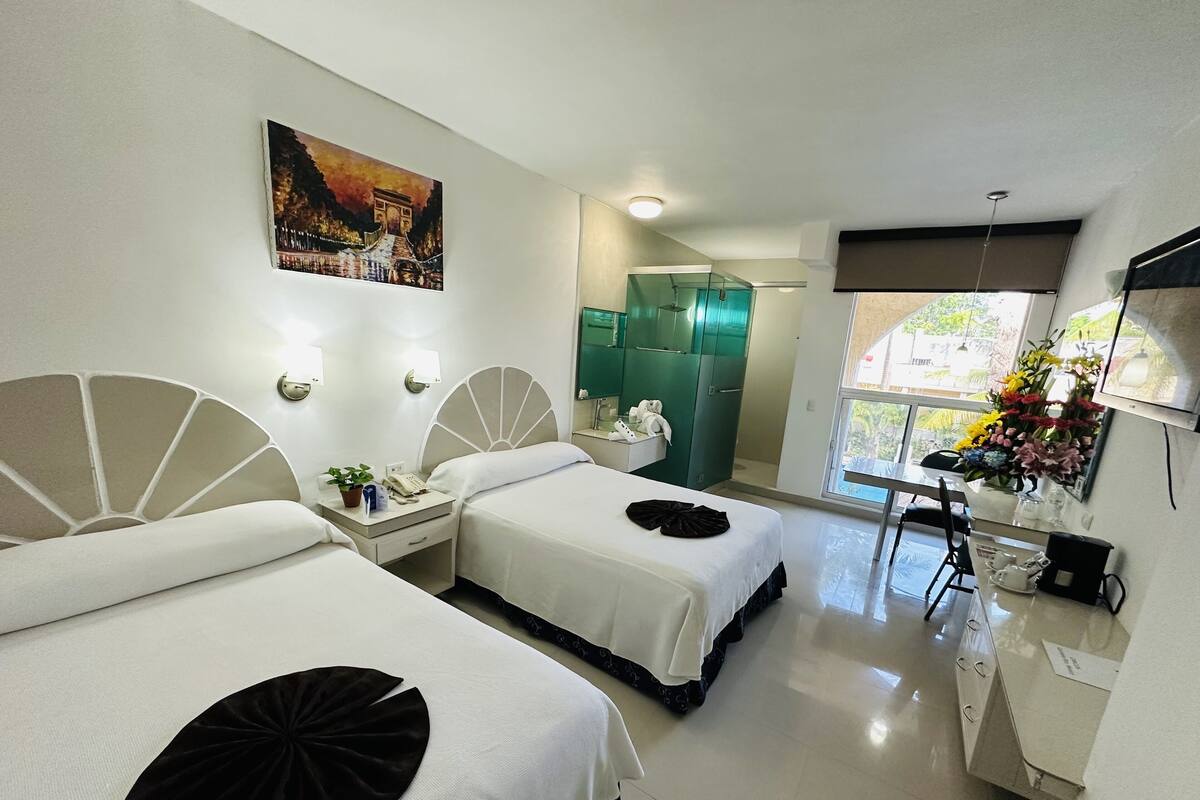 A hotel room with two beds, a bathroom with a glass shower, a desk with a chair, and a window with a view of the outside.