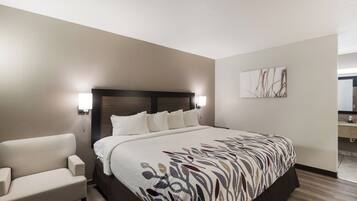 A hotel room with a large bed, a bedside table, a chair, a wall-mounted lamp, and a painting on the wall.