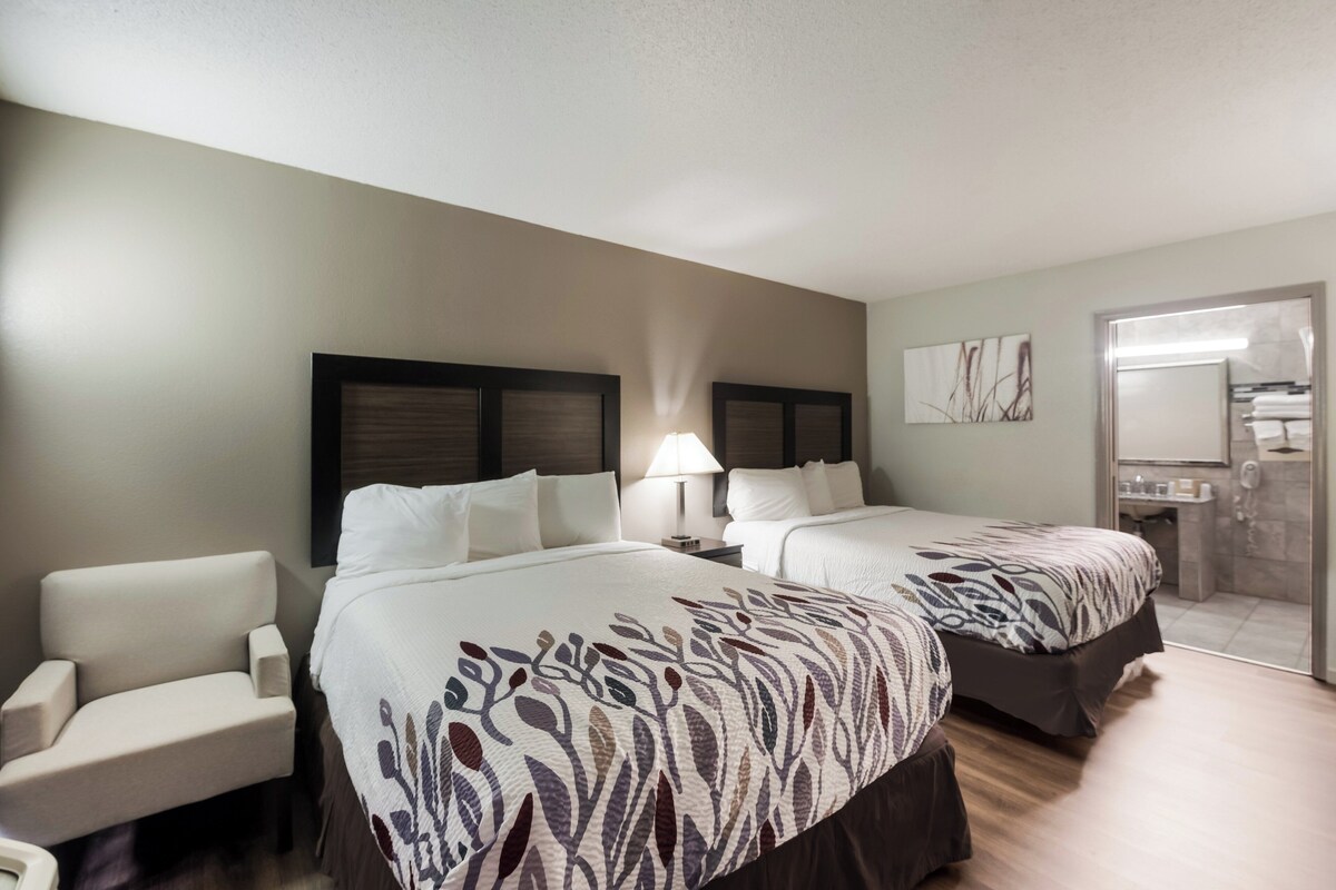 A hotel room with two beds, a chair, a nightstand, a lamp, and a bathroom visible through an open door.