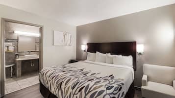A hotel room with a large bed, a bedside table, a chair, a bathroom with a sink and mirror, and a wall-mounted light fixture.