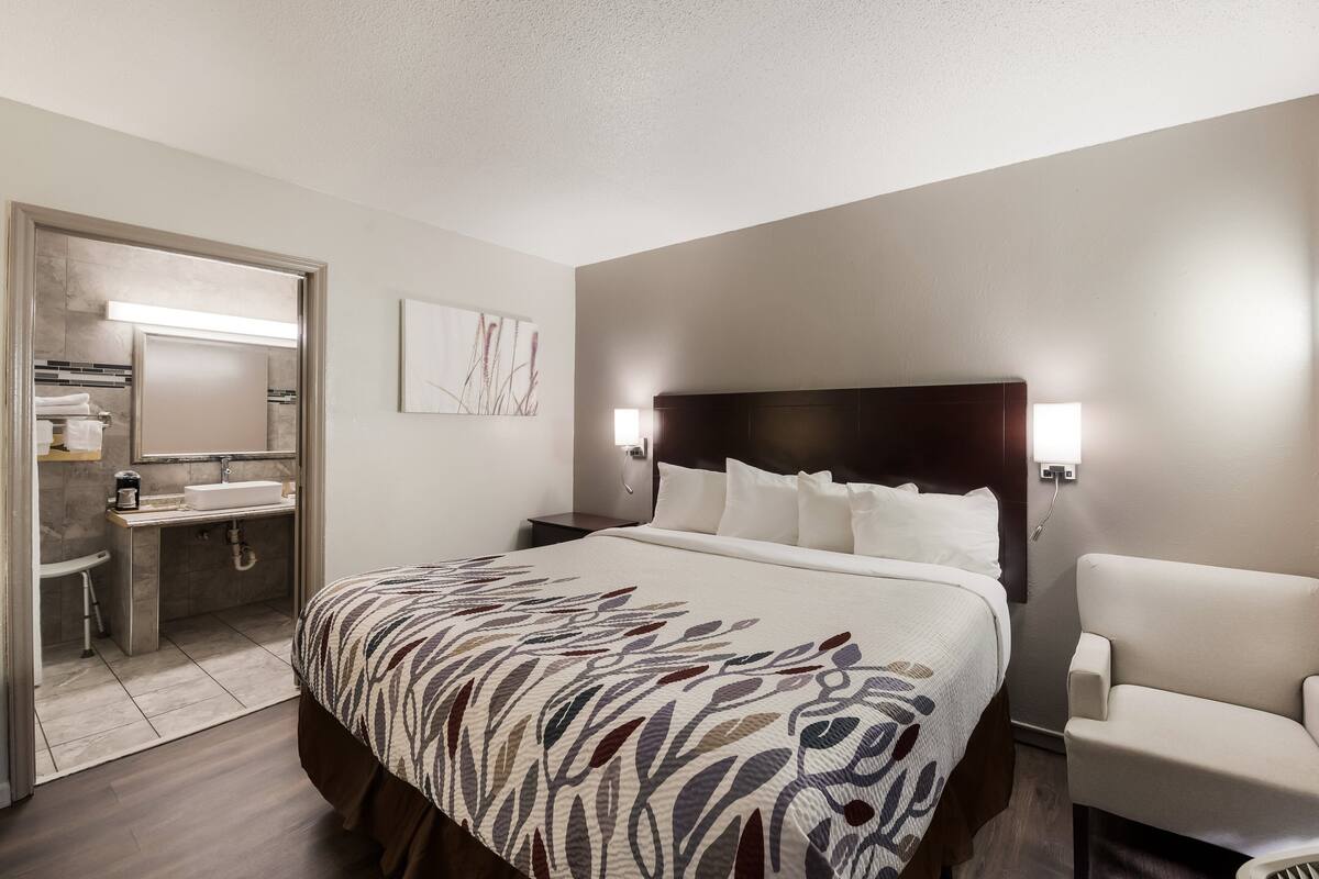 A hotel room with a large bed, a bedside table, a chair, a bathroom with a sink and mirror, and a wall-mounted light fixture.