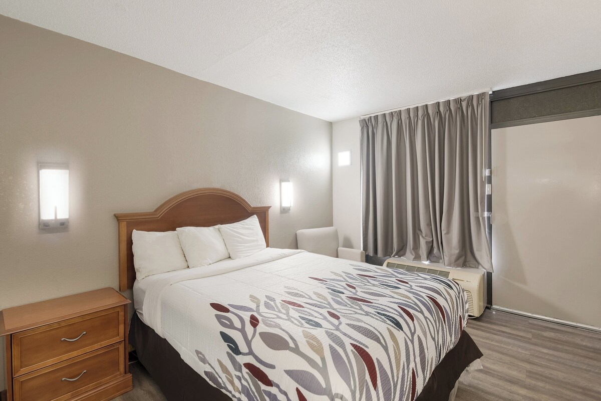 A hotel room with a large bed, a wooden nightstand, a chair, a window with curtains, and an air conditioning unit.