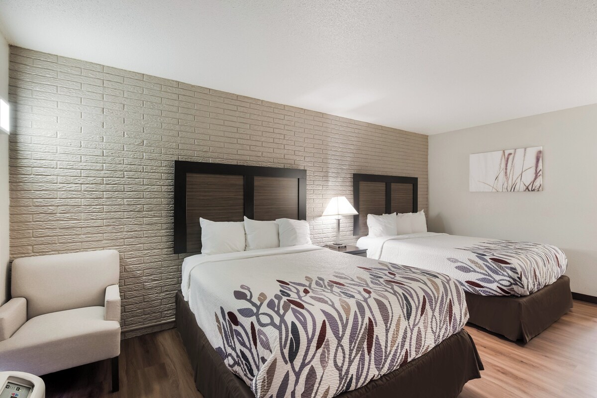 A hotel room with two beds, a brick-patterned wall, a wooden floor, a white armchair, and a nightstand with a lamp.