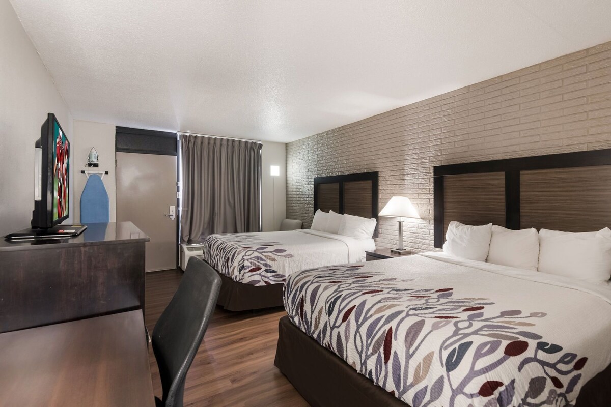 A hotel room with two beds, a desk, a TV, and a brick wall.