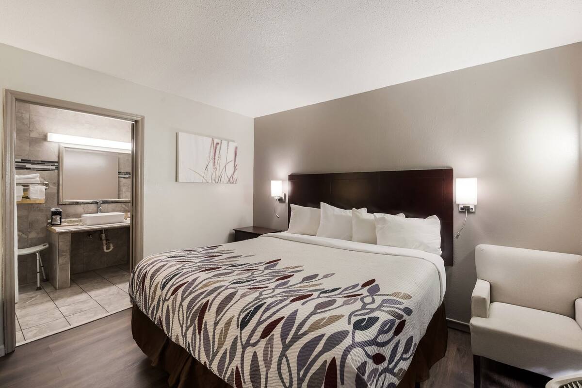 A hotel room with a bed, a chair, a nightstand, a wall-mounted lamp, and a bathroom visible through an open door.
