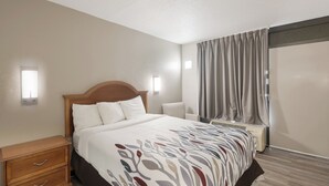 A hotel room with a bed, bedside tables, a nightstand, a wall-mounted light, a window with curtains, and a wall-mounted air conditioning unit. - Red Roof Inn Lenoir (Lenoir)