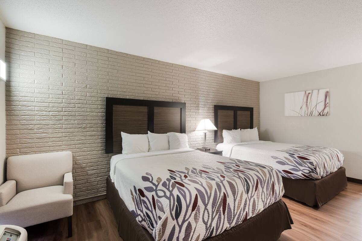 A hotel room with two beds, a brick-patterned wall, a wooden floor, a white armchair, and a nightstand with a lamp.