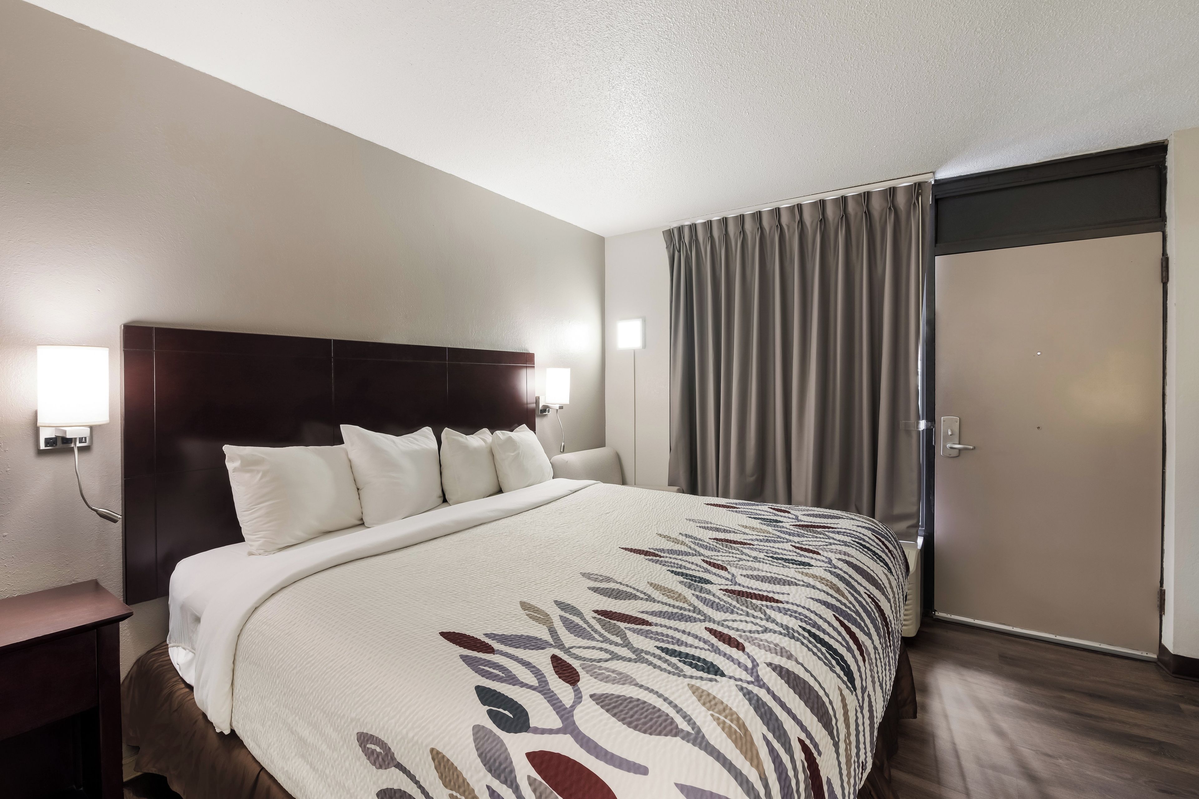 A hotel room with a large bed, a wooden headboard, a bedside table, a nightstand, and a window with curtains.