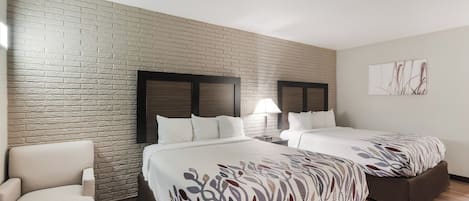 A hotel room with two beds, a brick-patterned wall, a wooden floor, a white armchair, and a nightstand with a lamp.