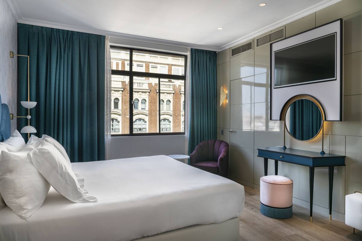 A modern hotel room with a large bed, a desk, a chair, and a view of a historic building.