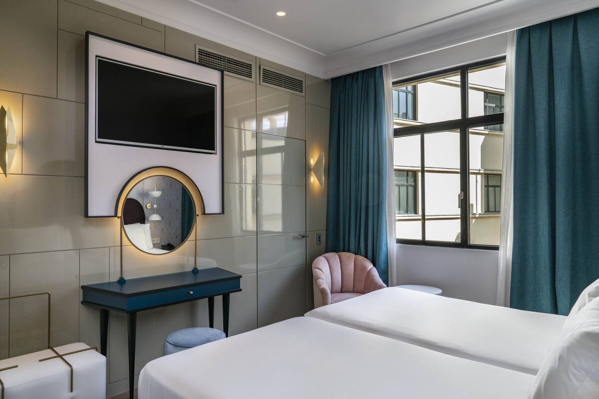 A hotel room with two beds, a desk with a mirror, a chair, and a large window with curtains.
